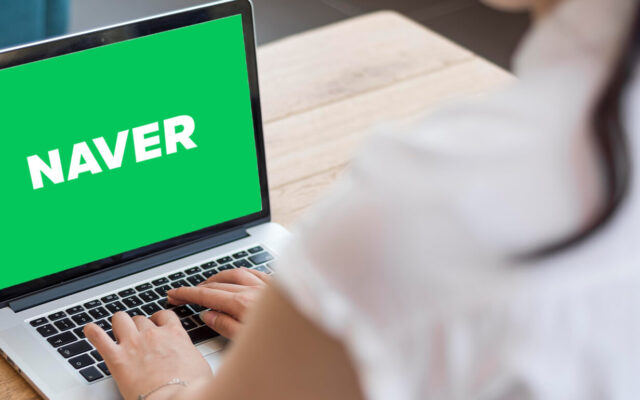 Person tippt auf einem Laptop mit grünem Bildschirm und weißem Schriftzug 'NAVER' auf einem Holztisch