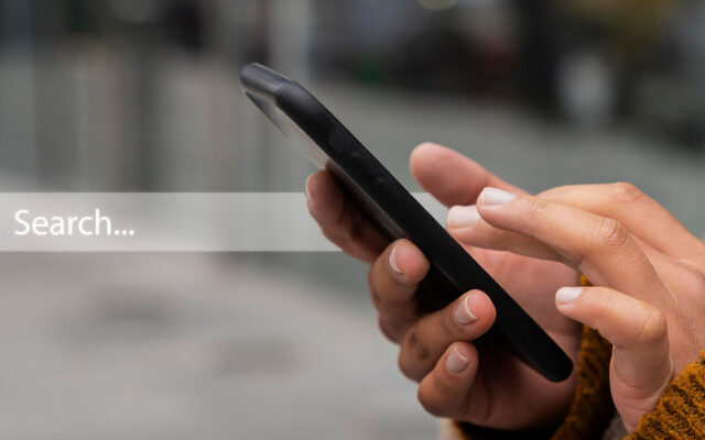 Hände einer Person mit braunem Strickpullover, die ein schwarzes Smartphone hält und darauf tippt, unscharfer Hintergrund.