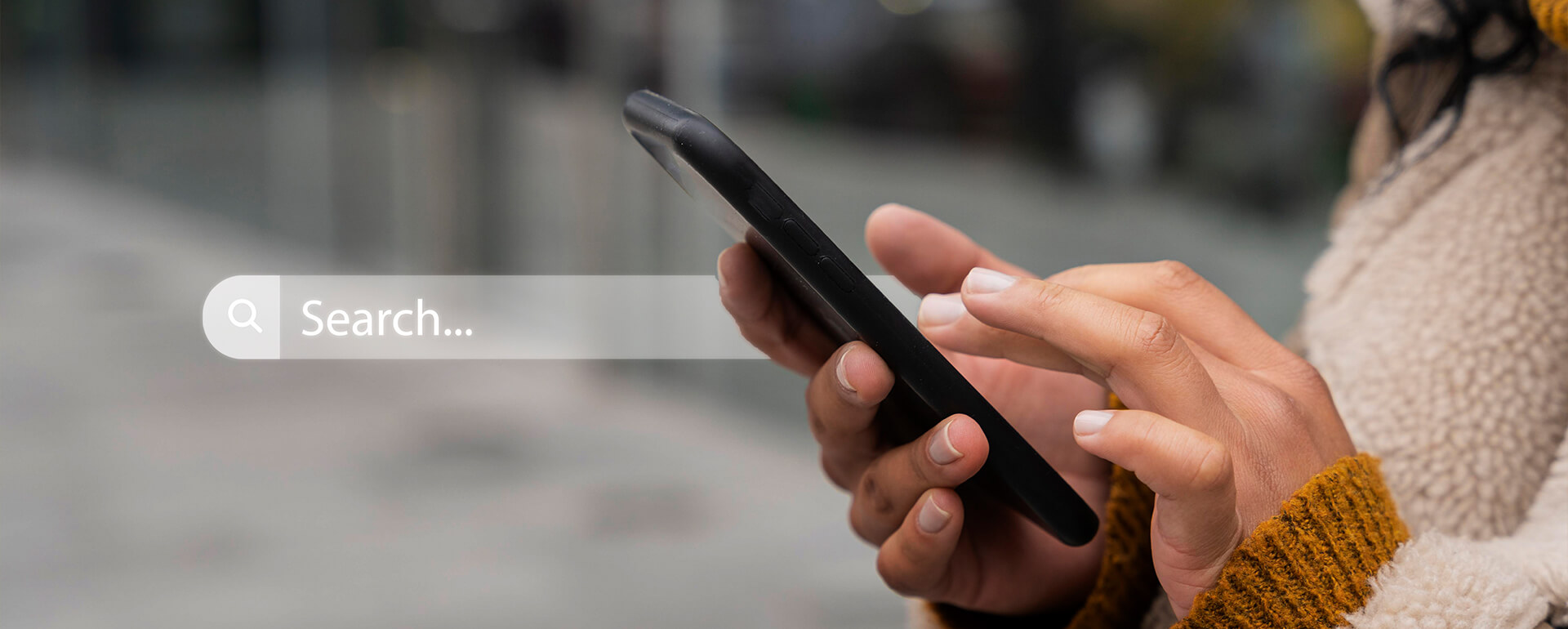 Hände einer Person mit braunem Strickpullover, die ein schwarzes Smartphone hält und darauf tippt, unscharfer Hintergrund.